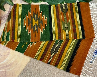 May include: Hand-woven textile with a geometric design. The rug features a central diamond pattern in shades of brown and yellow, surrounded by vertical stripes of green, brown, and gold. The edges have a fringe detail.