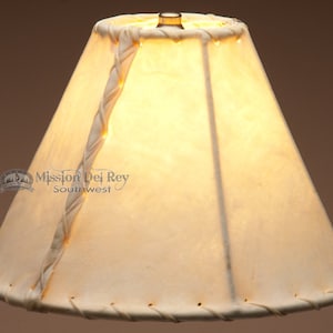 May include: A white leather lampshade with a braided leather trim. The lampshade is a cone shape and has a small metal top.