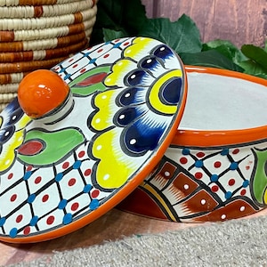 May include: A colorful ceramic dish with a lid. The dish has a white background with a floral pattern in shades of blue, yellow, green, and red. The lid has a bright orange knob.