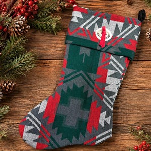 May include: A Christmas stocking featuring a woven geometric pattern in shades of dark green, red, gray, and white. The cuff is accented with a silver-toned metal bell tied with a thin red ribbon. A black fabric loop is attached to the top corner.