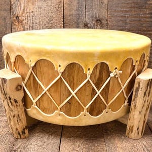 May include: A round drum with a light tan rawhide head stretched over a wooden frame. The head is secured with a white lacing pattern that creates a scalloped edge. The drum is supported by two thick, light-colored wooden legs with natural bark.