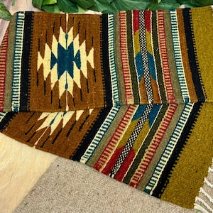 May include: A folded, rectangular woven textile with a geometric pattern. The design features a central diamond shape in teal and cream, surrounded by brown, black, and colorful stripes. The edges have a fringe detail.