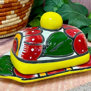 May include: A yellow ceramic butter dish with a red, green, and white floral design. The dish has a lid with a round knob.