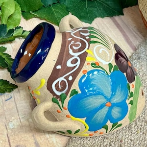 May include: Hand-painted ceramic mug with a blue rim and two handles. The mug features a large blue flower, brown and white decorative stripes, and green and yellow accents. A unique piece for beverages.
