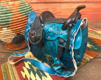 May include: A turquoise leather saddlebag with a horse design, black accents, and a braided strap. The bag features a saddle shape with a small pouch and is set on a colorful patterned textile.