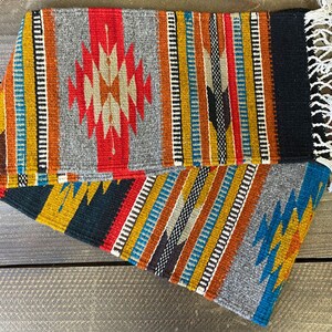 May include: A colorful woven rug with geometric patterns in shades of red, blue, yellow, and black. The rug has a fringe on one end and is made of wool.