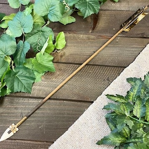 May include: A decorative arrow with a wooden shaft, a white arrowhead, and gold-colored fletching. The arrow is displayed on a wooden surface with green foliage.
