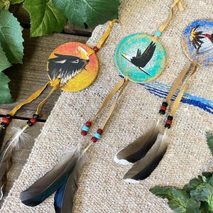 May include: Three circular hanging ornaments featuring hand-painted wildlife designs including a wolf, an eagle, and a hummingbird. Each ornament is suspended by a tan leather cord adorned with colorful beads and natural brown and grey feathers.