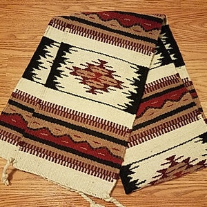 May include: A woven rug with a geometric pattern in shades of red, black, brown, and white. The rug is folded in half and has fringe on the ends.