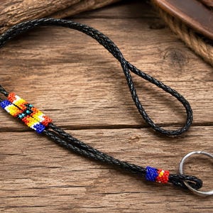 May include: A black braided leather lanyard featuring a rectangular beadwork section with red, yellow, orange, and blue geometric patterns, and a smaller matching beadwork accent near the silver metal key ring at the end of the cord.