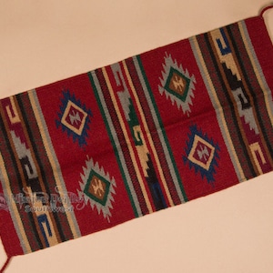 May include: A rectangular, woven rug with a red background and geometric patterns. The design includes diamond shapes and vertical stripes in shades of blue, green, brown, and tan. The rug has tassels at each end.