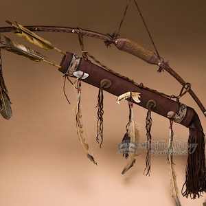 May include: A brown leather quiver with a wooden bow and three arrows. The quiver is decorated with feathers, beads, and a bone. The bow is strung and ready to shoot. The arrows are fletched with feathers. The quiver is hanging from a hook.