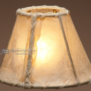 May include: A white leather lampshade with a braided leather trim. The lampshade is a cone shape and has a light bulb inside.