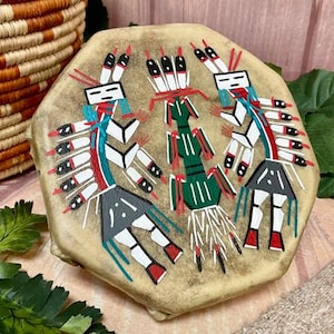 May include: Hand-painted Native American drum with a tan hide. The drum features three stylized figures in red, white, black, and green. A woven basket and greenery are in the background. The drum is a unique piece of art.