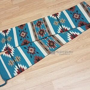 May include: A turquoise, white, brown, and red woven rug with a geometric pattern. The rug is folded in half and has fringe on the ends.
