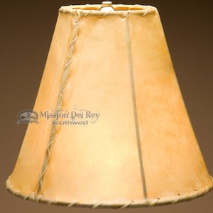 May include: A light brown leather lampshade with a braided leather trim. The lampshade is cone-shaped and has a subtle, rustic look.