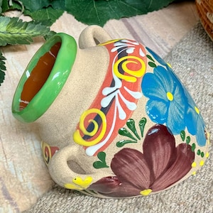 May include: A ceramic pitcher with a beige base, adorned with colorful floral patterns in blue, red, yellow, and maroon. The rim is a bright green, and it has two small handles.
