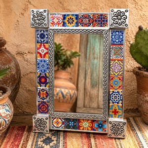 May include: A rectangular mirror featuring a silver metal frame decorated with colorful, patterned ceramic tiles in shades of blue, red, yellow, and orange. The four corners of the frame are accented with embossed silver floral metal squares.