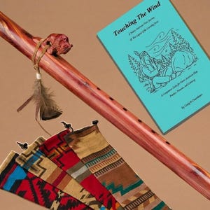 May include: A handcrafted wooden Native American flute with a reddish-brown finish, adorned with a carved bear and a feather. The image also shows a turquoise book titled "Touching The Wind" and several colorful woven flute bags.