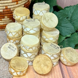 May include: A collection of small, round drums with tan drumheads and cream-colored sides. The sides are decorated with a geometric pattern. Some drums have "MADE IN MEXICO" stamped on them. The drums are stacked and arranged on a wooden surface.