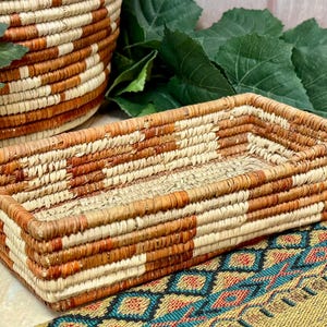 May include: A rectangular woven basket with a geometric pattern in shades of brown and cream. The basket is made of natural fibers and has a sturdy construction. A round basket is visible in the background.