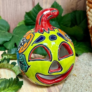 May include: A hand-painted ceramic pumpkin with a green base, red stem, and black eyes and mouth. The pumpkin is decorated with colorful floral patterns.