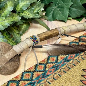 May include: A Native American ceremonial stone war club with a round stone head, wrapped in leather. The wooden handle is adorned with colorful beads and a feather. The club rests on a patterned textile.