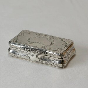 May include: A silver rectangular box with an ornate engraved design. The box has a hinged lid and is decorated with intricate floral patterns.