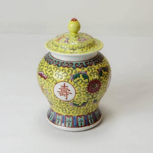 May include: A yellow porcelain jar with a lid, decorated with floral patterns and Chinese characters. The jar has a gold trim and a blue and green base.