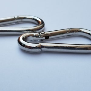 May include: Two silver metal snap hooks, also known as carabiners, are shown on a white background. The hooks are closed and lying side by side.