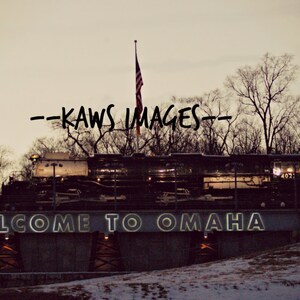 May include: A black train engine with a sign that reads "WELCOME TO OMAHA" in front of it. The train is parked in front of a building with trees in the background. The sky is a light blue color.
