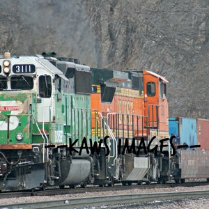 May include: A green and white train engine with the number 3111 on the side, pulling a string of orange and blue freight cars. The train is on a set of tracks with a snowy background.