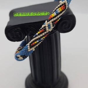 May include: A beaded bracelet with a blue background and a geometric pattern in white, red, yellow, and black. The bracelet is displayed on a black decorative column. The text "BEADEDIEZ" is visible.