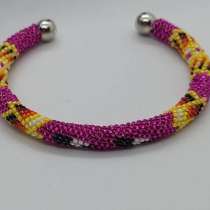May include: A beaded cuff bracelet in shades of fuchsia, yellow, orange, and black. The bracelet has a repeating pattern of colors and is finished with silver-colored metal ball ends.