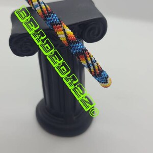May include: A colorful, beaded bracelet in shades of blue, yellow, orange, and black rests on a black decorative column. The bracelet is cylindrical and features a detailed, woven pattern. The image also includes the text "BEARDEDMAN" in green.