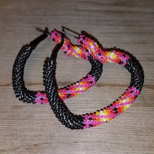 May include: A pair of heart-shaped hoop earrings. The lower half of each earring is black, while the upper half features a pattern of pink, yellow, and white beads. The earrings are displayed on a wooden surface.