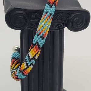 May include: A colorful beaded bracelet with a spiral design, featuring turquoise, yellow, orange, red, and black beads. The bracelet is displayed on a black decorative column. The bracelet has a silver clasp.