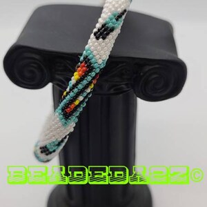 May include: A beaded bracelet with a white base, adorned with turquoise, black, orange, and yellow beads in a geometric pattern. The bracelet is displayed on a black pedestal. The text "BEADED22" is visible.