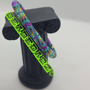 May include: A colorful beaded bracelet with a mix of purple, teal, yellow, and orange beads. The bracelet is displayed on a black decorative pillar. The image also contains the text "BEADED DREAM" in neon green.