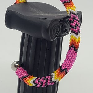 May include: A beaded bracelet with a vibrant pattern of pink, black, yellow, orange, and white beads. The bracelet is displayed on a black pedestal and has a silver magnetic clasp.