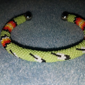 May include: A beaded bracelet with a light green base color, accented with black, white, red, orange, and yellow beads. The bracelet has a spiral design and features silver metal end caps. The bracelet is a piece of jewelry.