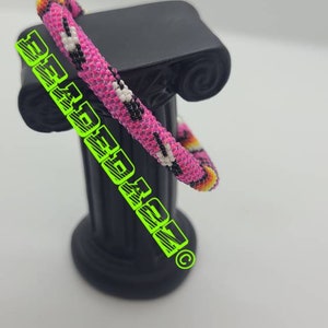 May include: A beaded bracelet with a pink base, featuring a pattern of black and white spots. The bracelet is displayed on a black pedestal. The text "BEADEDAZN" is overlaid on the image in green.
