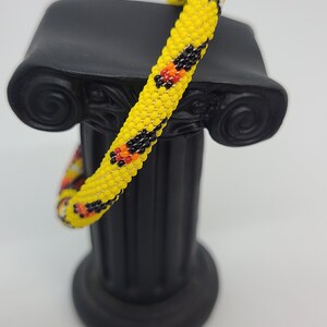 May include: A beaded bracelet with a yellow base color, accented with black, red, and orange patterns. The bracelet is displayed on a black decorative column against a white background. The bracelet is a piece of jewelry.