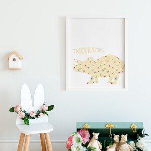 May include: A framed print featuring a yellow triceratops silhouette filled with floral patterns. The word "TRICERATOPS" is printed above the dinosaur. A white bunny-eared chair with pink flowers and a wooden birdhouse are also visible.