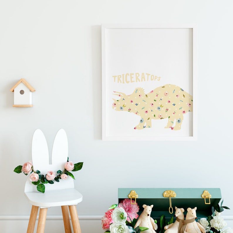 May include: A framed print featuring a yellow triceratops silhouette filled with floral patterns. The word "TRICERATOPS" is printed above the dinosaur. A white bunny-eared chair with pink flowers and a wooden birdhouse are also visible.