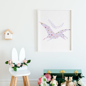 May include: A framed print featuring a lavender Pterodactyl silhouette filled with a floral pattern. A white bunny-eared chair is decorated with pink roses and greenery. A small wooden birdhouse and a teal chest with gold hardware complete the scene.