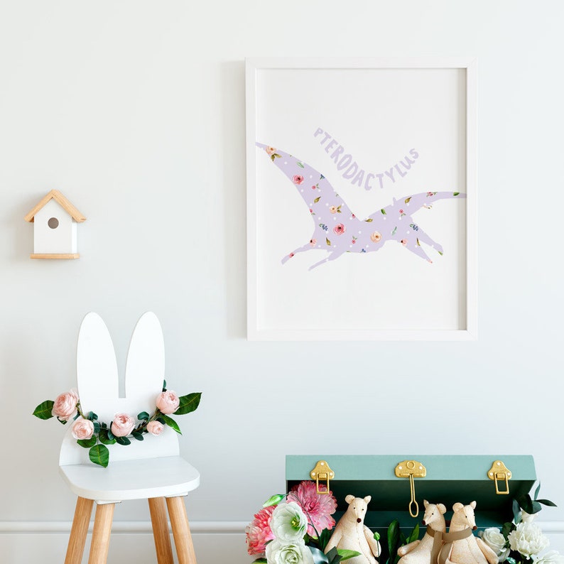 May include: A framed print featuring a lavender Pterodactyl silhouette filled with a floral pattern. A white bunny-eared chair is decorated with pink roses and greenery. A small wooden birdhouse and a teal chest with gold hardware complete the scene.