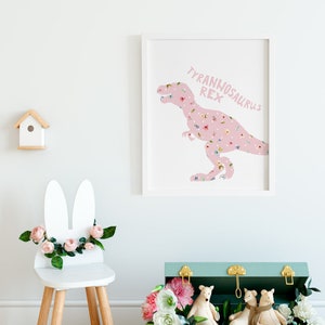 May include: A framed print featuring a pink Tyrannosaurus Rex silhouette filled with a floral pattern. The text "TYRANNOSAURUS REX" is above the dinosaur. A white chair with bunny ears and flowers, a birdhouse, and a teal chest with toys are also in the image.