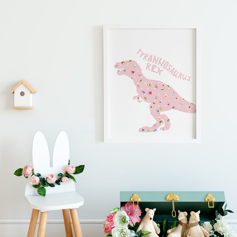 May include: A framed print featuring a pink Tyrannosaurus Rex silhouette filled with a floral pattern. The text "TYRANNOSAURUS REX" is above the dinosaur. A white chair with bunny ears and flowers, a birdhouse, and a teal chest with toys are also in the image.