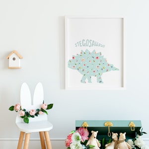 May include: A framed print featuring a light blue Stegosaurus dinosaur silhouette filled with colorful flowers and white polka dots. A white bunny-eared chair with pink flowers and a small wooden birdhouse are also visible.